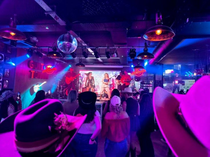 View of back of crowd wearing cowboy hats, watching a 5-piece band on stage