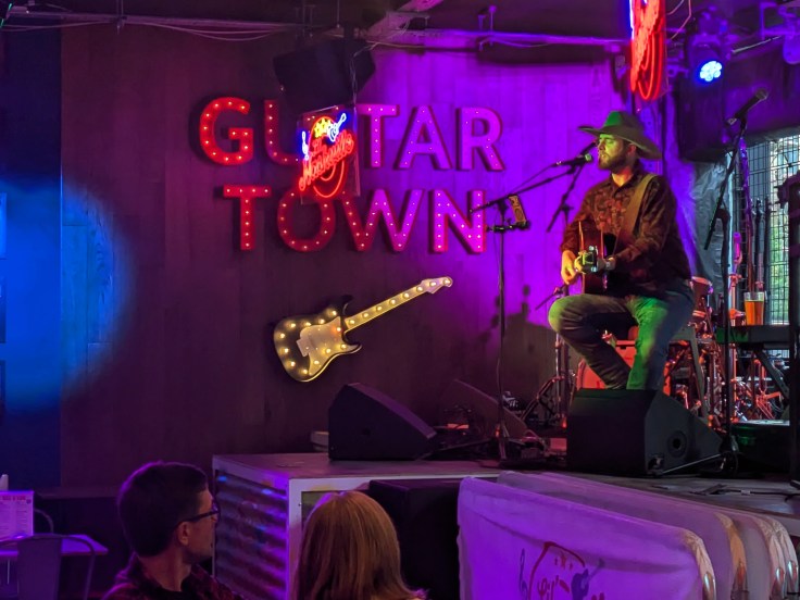Singer sat on a stool on the Lil Nashville bar stage. He is wearing a cowboy hat and holding a guitar.